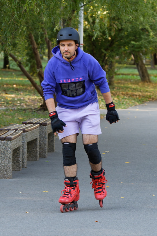 10 - A man inline skating along an outdoor path while wearing a helmet, wrist guards, knee pads, a purple hoodie, and bright red inline skates